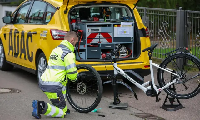 ADAC-Mitarbeiter repariert eine Fahrradpanne. Ein gelbes ADAC-Auto mit Werkzeugkasten steht daneben.