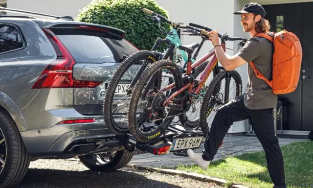 Mężczyzna z pomarańczowym plecakiem montuje bagażnik rowerowy na hak do Volvo XC60. Dwa rowery górskie są już zamocowane. Przepisy dotyczące bagażnika rowerowego na hak są kluczowe dla bezpieczeństwa.