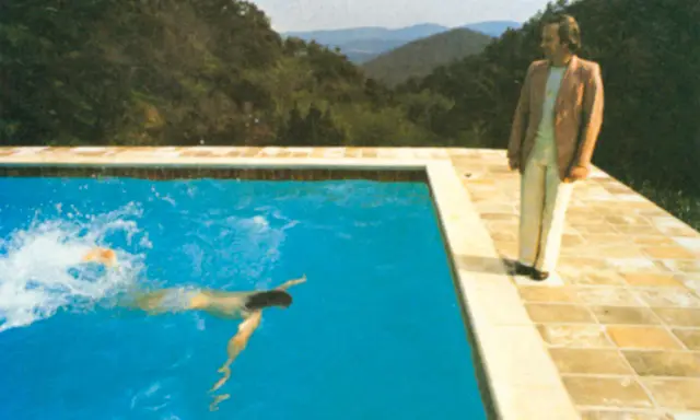 A portrait of an artist by a pool with two figures. One figure swims in the bright blue water, while another stands on the tiled edge, looking out at the mountainous landscape.