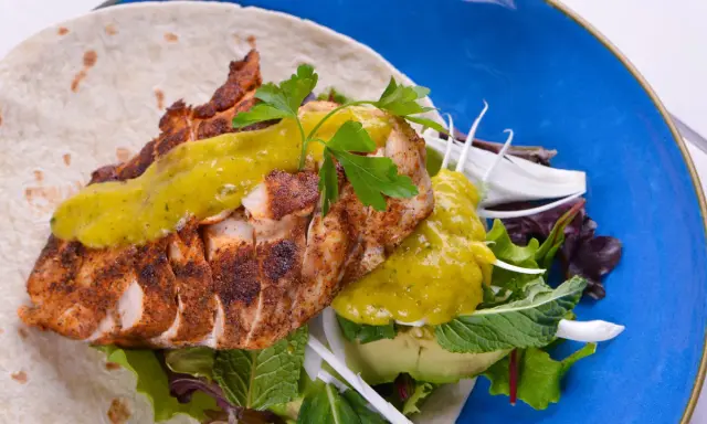 Delicioso wrap de pollo con aguacate, lechugas variadas y salsa verde, servido en un plato azul vibrante.