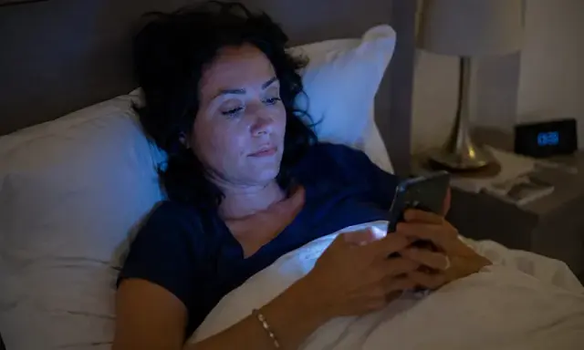 Une femme dans son lit, éclairée par la lumière bleue de son téléphone, perturbant son sommeil.