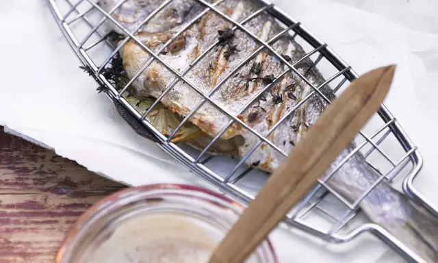 Ein ganzer Fisch liegt in einem Grillkorb, bereit zum Fisch grillen. Zitronenscheiben und Kräuter sind darin versteckt.