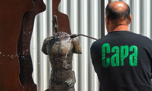 Hombre con camiseta negra con letras verdes "Capa" limpia una estatua de bronce con una manguera a presión.