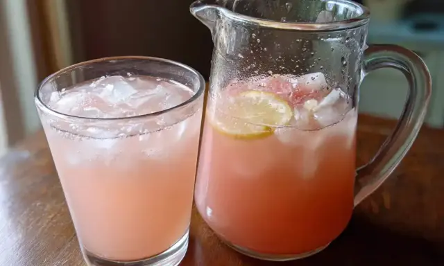 Ett glas och en karaff fyllda med is och en rosa rabarbersirap drink. Citronskivor flyter i karaffen.