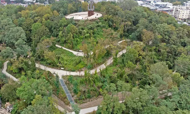 Wzgórze z pomnikiem Kotwicy, otoczone zielenią. Kręte ścieżki i schody prowadzą przez park akcji "Burza", oferując widok na miasto i Wisłę.