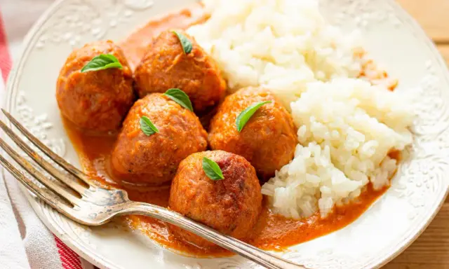 Albondigas a la jardinera con arroz blanco, decoradas con hojas verdes. Un tenedor plateado descansa sobre el plato.