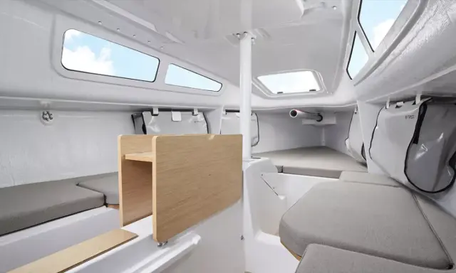 Intérieur du FIRST 24, cabine lumineuse avec banquettes grises, table en bois et hublots ouverts sur le ciel bleu.