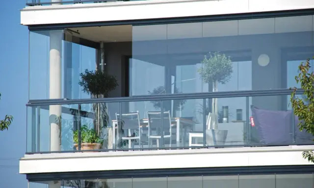 Un balcone moderno con vetrate scorrevoli, ideale per chiudere un balcone fai da te. Tavolo, sedie e un pouf viola creano un angolo relax.