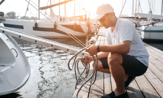 Un homme prépare une corde sur un ponton, près d'un bateau. Il manipule le cordage, peut-être pour un système de poulie.