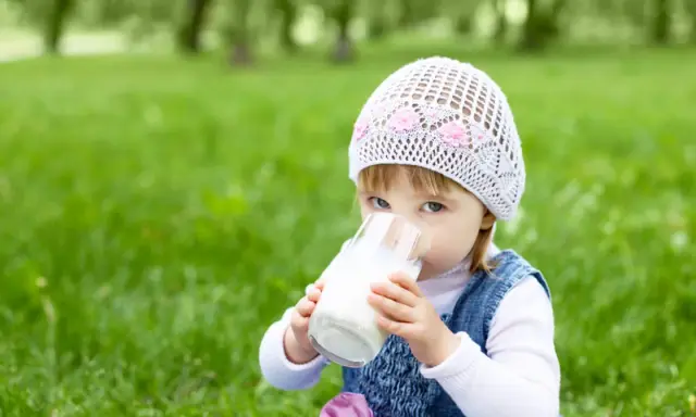 Mała dziewczynka w czapce pije mleko owsiane dla niemowlaka ze szklanki na trawie.