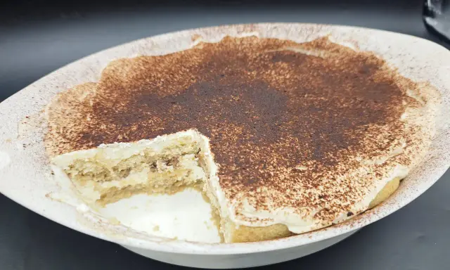 Un délicieux tiramisu sans oeuf, crémeux et saupoudré de cacao, dans un plat blanc. Une part a été retirée, révélant les couches de biscuits et de crème.