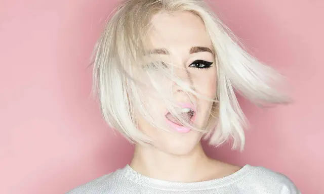 Mujer con cabello rubio platino y corte recto al hombro, con un ojo visible y labios rosados abiertos.