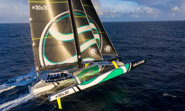 Un trimaran aux voiles noires et blanches, arborant le logo "sails of change", fend les vagues bleues sous un ciel nuageux.
