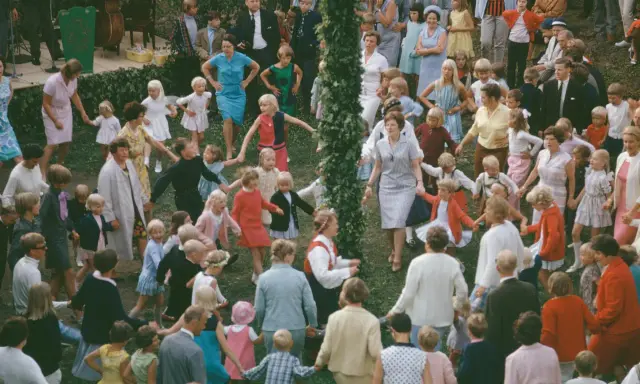 Många barn och vuxna dansar ringdans runt en midsommarstång i Sverige.