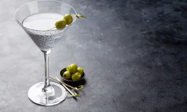 Ein Glas trockener Wermut mit Oliven garniert, daneben eine Schale mit weiteren Oliven auf dunklem Untergrund.
