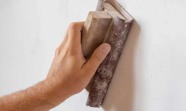 Mano lijando paredes para pintar. El bloque de madera con lija se desliza sobre la superficie blanca, preparándola para un acabado perfecto.