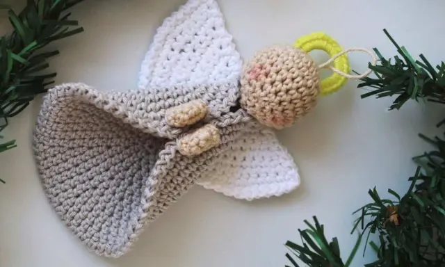 Un tierno angel amigurumi de ganchillo, con alas blancas y un cuerpo gris, adornado con ramitas verdes.