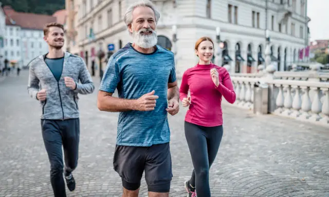 Un uomo con barba bianca e due giovani corrono sorridenti in città. Un esempio di come iniziare a correre a 50 anni sia possibile e gratificante.