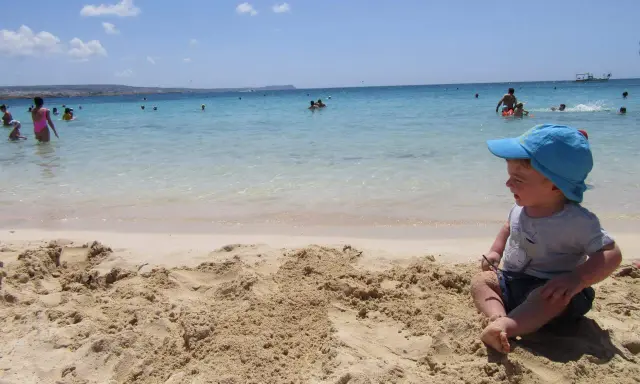 Bambino con cappellino blu gioca sulla sabbia a Cipro, mentre altri si divertono in mare.