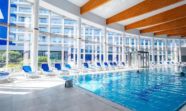 Wnętrze z basenami hotelowymi, leżakami i dużymi oknami z widokiem na budynek hotelowy.