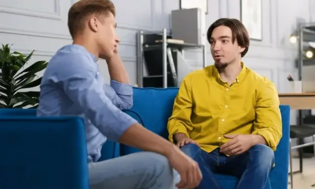 Dwóch mężczyzn siedzi na kanapie, rozmawiając. Jeden z nich opowiada o tym, jak przełamać barierę językową, szukając inspiracji w szczerej rozmowie.