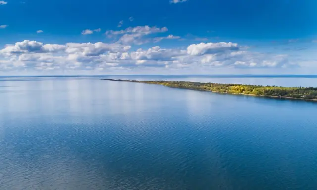 Blå himmel med moln över ett lugnt vatten. En lång, grönskande udde sträcker sig ut. Perfekt för vandring Mariestad.