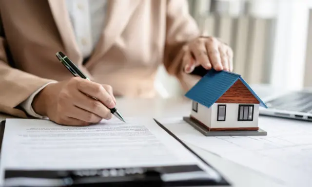 Ręka podpisuje dokumenty przy makiecie domu. Unikaj problemów, kiedy nie płaci się PCC od zakupu mieszkania.