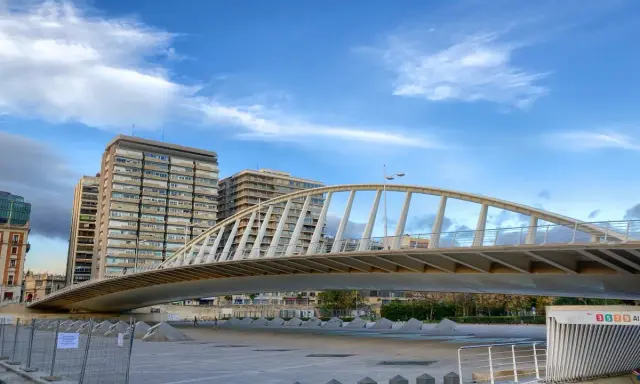 El moderno puente de la Exposición cruza un río, con edificios altos al fondo bajo un cielo azul con nubes.