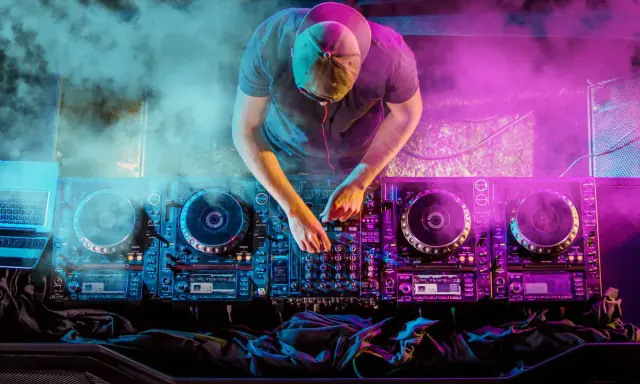 DJ con cuffie e cappellino mixa musica hause su console illuminata da luci viola e blu, circondato da fumo.