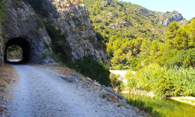 Camino de grava junto a un río y un túnel en la vía verde de Alicante. Naturaleza exuberante y rocas escarpadas.