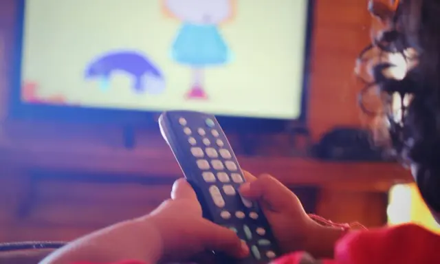Dziecko z pilotem w ręku ogląda bajkę na telewizorze w pokoju.