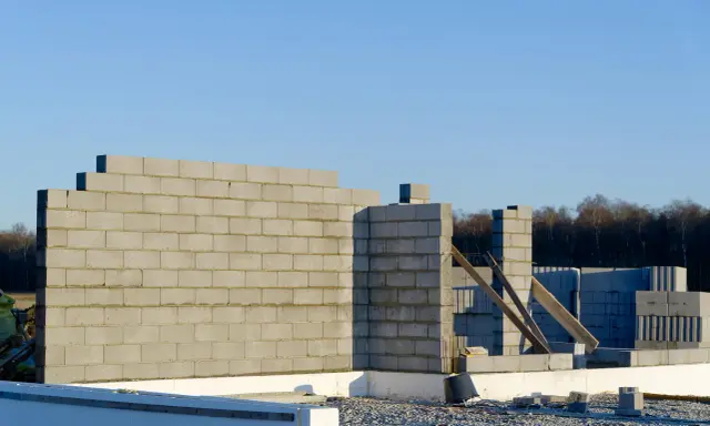 Budowa domu w toku. Wznoszona jest ściana nośna z bloczków betonowych, widoczne fundamenty i materiały budowlane.