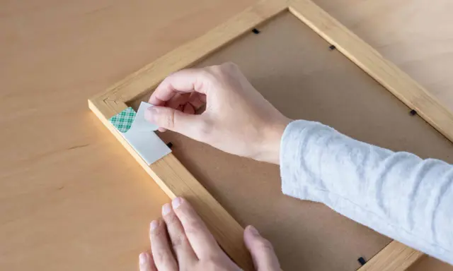 Mano retirando cinta de doble cara de un marco de madera. Aprende cómo quitar cinta de doble cara de la pared sin dañarla.