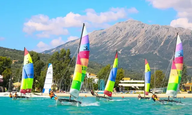 Plusieurs Hobie Cat 16 naviguent sur une mer turquoise, voiles multicolores, montagne en arrière-plan.
