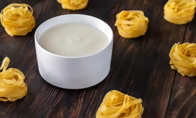 Un bol de béchamel sans beurre crémeuse, entouré de nids de pâtes fraîches sur fond de bois sombre.