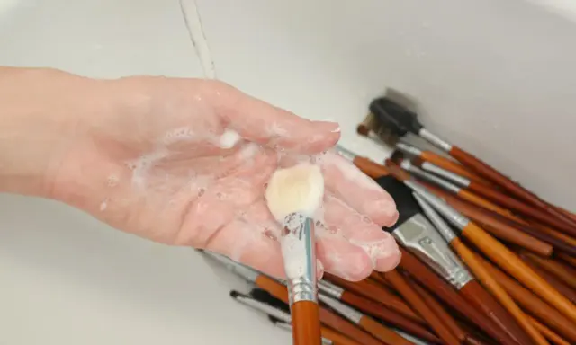 Mano con espuma jabonosa limpiando brochas de maquillaje en un lavabo.