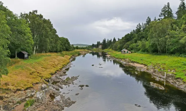Ein ruhiger Fluss, der durch eine grüne Landschaft fließt. Kleine Häuser säumen das Ufer, vielleicht Orte, an denen man das schottische Uisge Beatha genießt.