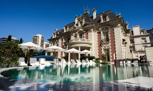 En lyxig pool framför en ståtlig byggnad som påminner om White Lotus Hotel, med solstolar och parasoller.