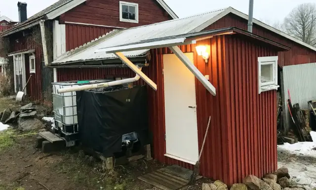 Rött utedass med vit dörr och fönster. Bredvid står en stor tank och en svart presenning. Här följer man utedass regler.