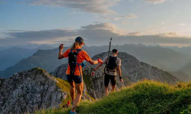 Dwie osoby w sportowym stroju, z plecakami i kijkami, biegną po górskim szlaku. Idealny przykład, jak się ubrać w góry na bieg.