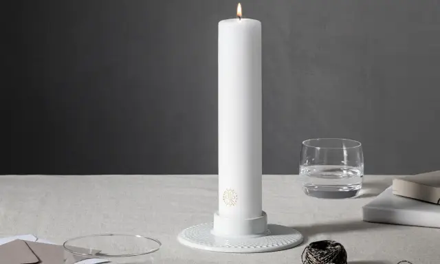 Ett vitt, högt stearinljus med guldig logotyp står i en vit ljusstake. Bredvid finns ett glas vatten, böcker och garn. Olika former av stearinljus kan skapa stämning.