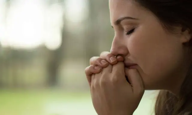 Visage pensif d'une jeune femme, mains jointes devant sa bouche, peut-être réfléchissant aux tactiques d'un manipulateur narcissique.