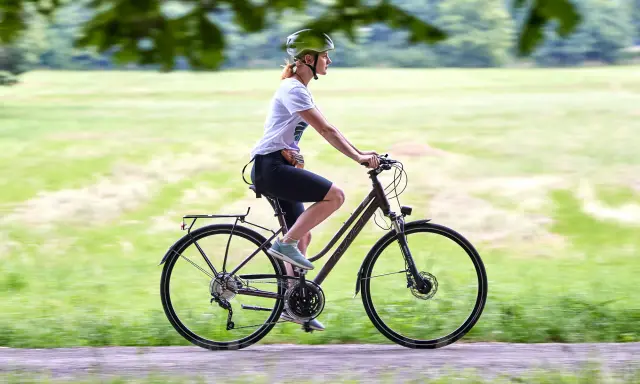 Kobieta w kasku jedzie na rowerze, który może być zarówno rowerem crossowym, jak i trekkingowym, po ścieżce w parku.