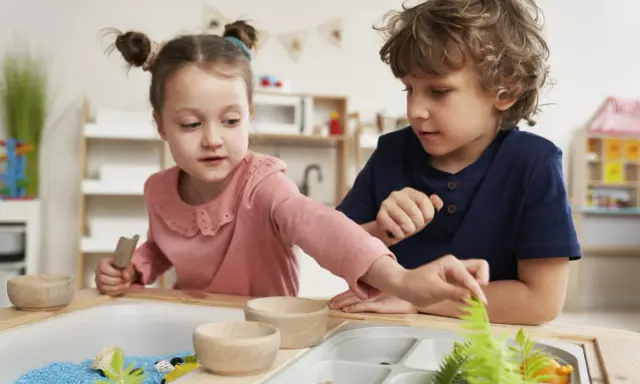 Dwoje dzieci bawi się w piaskownicy sensorycznej z niebieskim piaskiem i roślinami.