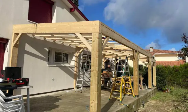 Construction d'une charpente sans ferme en bois, avec des poteaux et des poutres, sur une terrasse. Des échelles sont présentes.