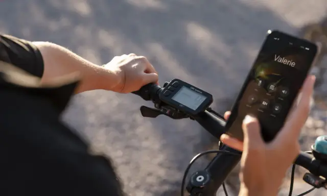 Rowerzysta trzyma telefon, na którym wyświetla się połączenie z Valerie. Obok widoczne są symbole na liczniku rowerowym, pokazujące prędkość i inne dane.