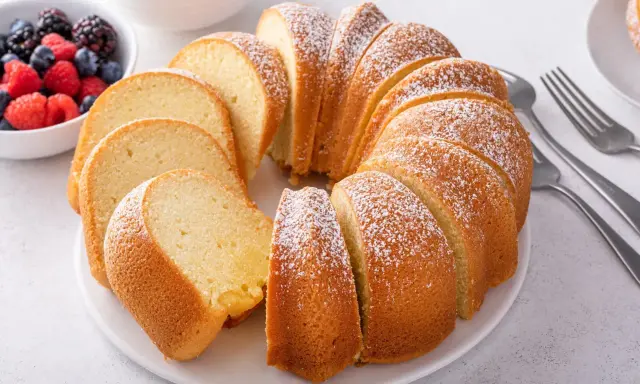 Pokrojona babka piaskowa, oprószona cukrem pudrem, z miseczką owoców leśnych w tle.