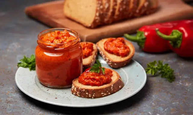 Słoik pełen ajvaru, pysznej pasty z papryki, podany z chlebem i świeżymi paprykami. Ajvar co to? To smak Bałkanów!