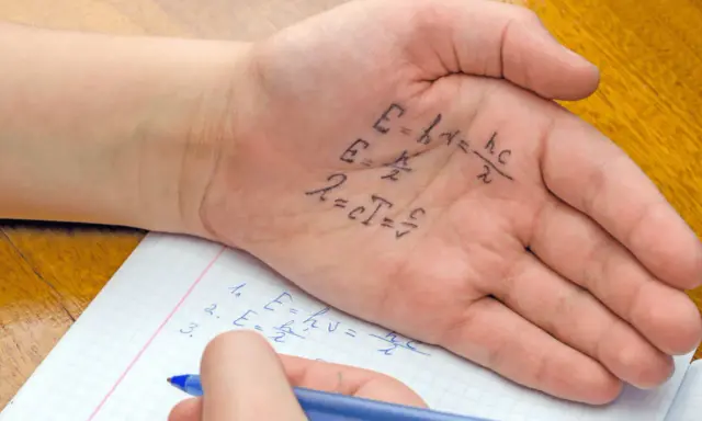 Formeln auf der Handfläche, um in der Schule zu spicken. Ein Schüler schreibt physikalische Gleichungen auf seine Hand, während er in sein Heft schaut.