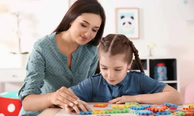 Kobieta pomaga dziewczynce z niepełnosprawnością intelektualną w stopniu lekkim układać kolorowe puzzle.
